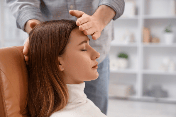 A woman sitting on a sofa with her eyes closed, whilst a man therapist conducts a hypnotherapy session.
